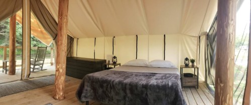Cozy glamping tent interior with a bed, wooden furniture, and lanterns, surrounded by nature.