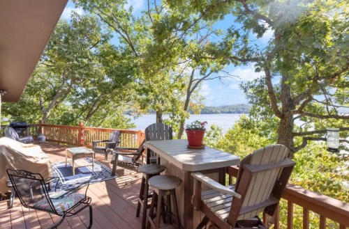 A scenic deck with wooden furniture, overlooking a lake surrounded by trees under a bright blue sky.