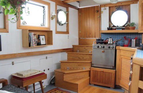 Cozy kitchen area with wooden stairs, porthole windows, and a stove, featuring plants and warm wood accents.