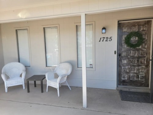 A porch with two white chairs and a small table, featuring a wooden door with a wreath and house number 1725.