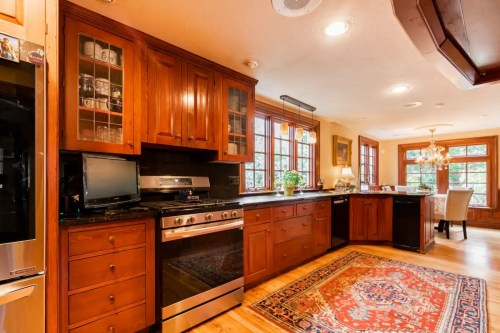 A warm, wooden kitchen with a large stove, dark countertops, and a colorful rug, featuring windows and a dining area.