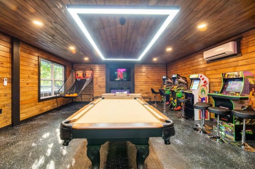 A game room featuring a pool table, arcade machines, and wooden walls, illuminated by a modern ceiling light.