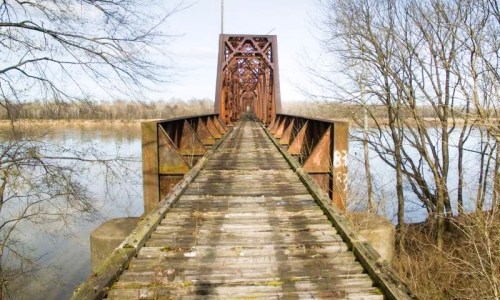 Rusty train bridge extending over a calm river, surrounded by bare trees and a clear blue sky.