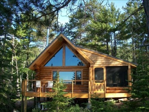 A cozy wooden cabin with large windows, surrounded by tall trees and greenery, featuring a deck and a sloped roof.