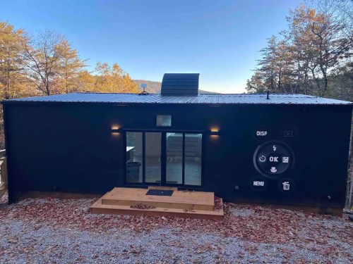 A modern black house with a wooden deck, surrounded by autumn trees and a clear blue sky.