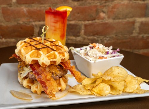A waffle sandwich with a burger, bacon, and onions, served with chips and coleslaw, alongside a colorful drink.