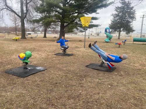 Two children play on spring riders in a park, with trees and playground equipment in the background.