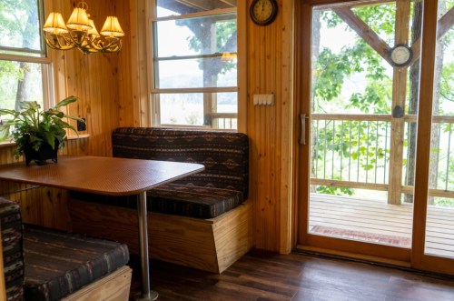 Cozy wooden interior with a dining table, benches, and a door leading to a deck surrounded by greenery.