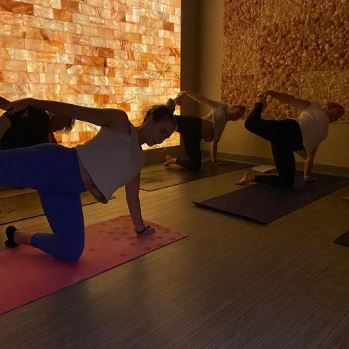 A group of people practicing yoga in a dimly lit studio with a textured wall in the background.