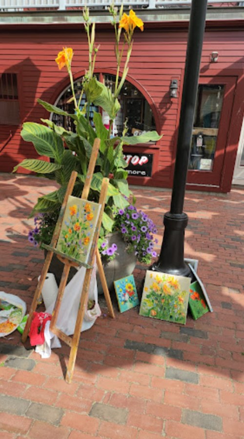 An easel with flower paintings beside a large planter and colorful flowers on a brick sidewalk.