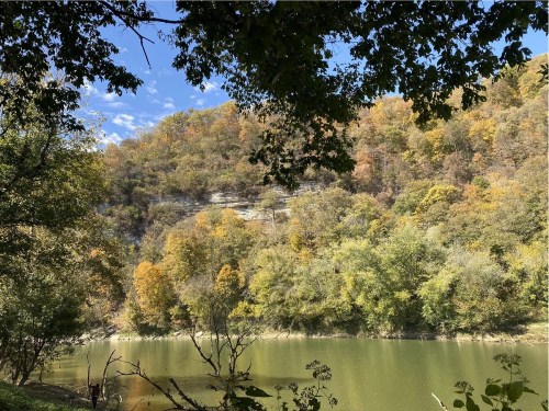 A serene river scene framed by trees, with a hillside displaying autumn foliage in warm colors.