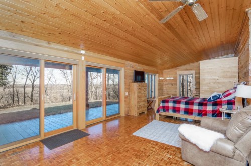 Cozy wooden cabin interior with a plaid bedspread, large windows, and a scenic view of nature outside.
