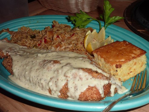 A plate featuring fried fish with creamy sauce, rice, lemon wedges, and a piece of cornbread, garnished with parsley.