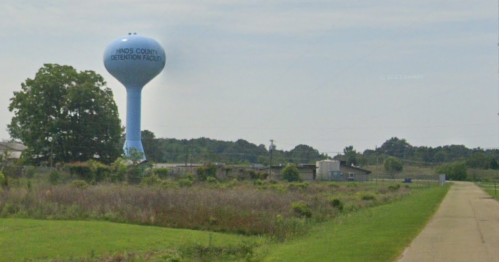 A blue water tower stands near a detention facility, surrounded by grassy fields and a dirt path.