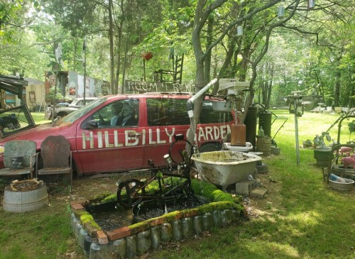 A red van labeled "Hillbilly Garden" surrounded by eclectic yard art and greenery in a rustic outdoor setting.