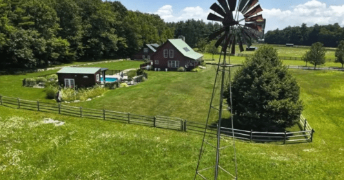 Aerial view of a green landscape featuring a house, windmill, pool, and trees under a blue sky.