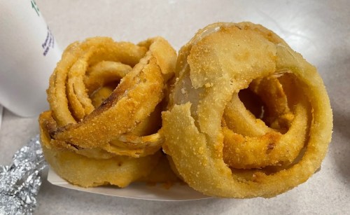 Two crispy, golden-brown onion rings stacked on a white tray, with a soft drink in the background.