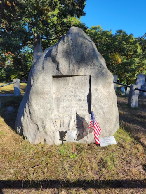 A large rock grave marker with an engraved plaque, surrounded by trees and a small American flag.