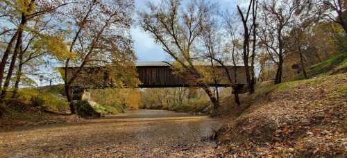 A covered bridge spans a dry riverbed, surrounded by autumn trees with colorful leaves.