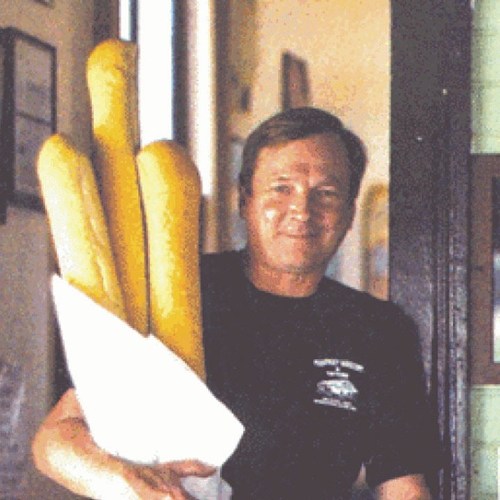 A man smiles while holding a large bundle of fresh baguettes wrapped in paper.