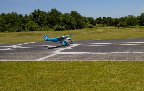 A small blue airplane takes off from a grassy runway on a sunny day, surrounded by trees.