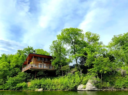A wooden house perched on a rocky hillside, surrounded by lush green trees and a calm body of water below.