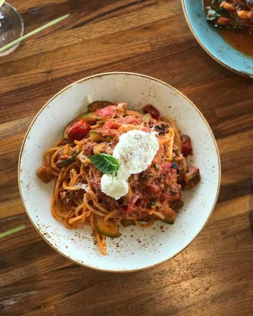 A plate of spaghetti topped with tomato sauce, vegetables, and a dollop of ricotta cheese, garnished with a basil leaf.