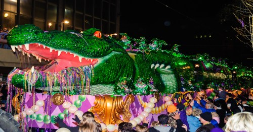 A large, green alligator float adorned with beads and decorations moves through a nighttime parade, surrounded by cheering crowds.