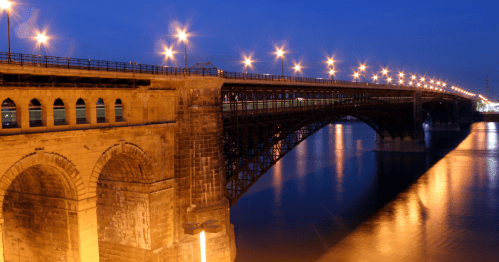 A bridge illuminated by streetlights spans a calm river at dusk, reflecting warm light on the water's surface.