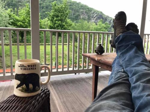 A person relaxes on a porch with feet up, a coffee mug, and a scenic view of greenery and mountains.