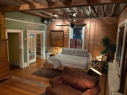 Cozy bedroom with exposed beams, a bed, a sofa, and warm lighting, featuring brick walls and wooden floors.
