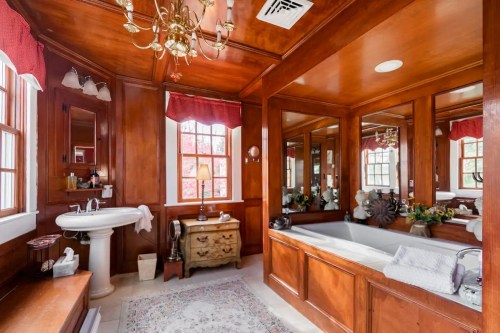 A warm wooden bathroom featuring a bathtub, sink, chandelier, and large mirrors, with natural light from windows.