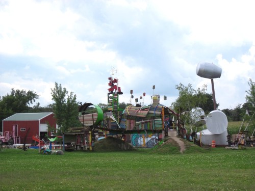 Colorful, whimsical playground structure with various shapes and designs, set against a cloudy sky and green grass.