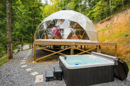 A geodesic dome cabin surrounded by trees, featuring a hot tub on a gravel path.