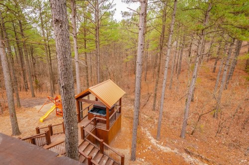 A wooden play structure with a slide surrounded by tall pine trees and autumn foliage.