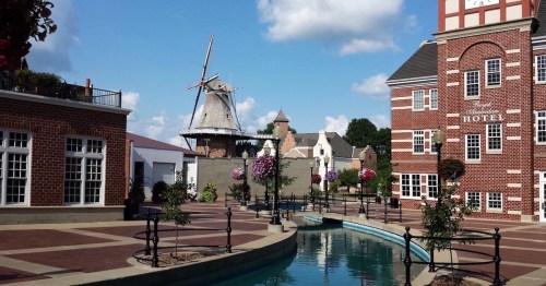 A scenic view of a canal with flowers, a windmill, and brick buildings under a blue sky with fluffy clouds.