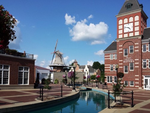 A charming plaza with a clock tower, windmill, and a canal, surrounded by flowers and blue skies.