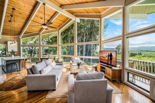 Bright, modern living room with large windows, wooden ceiling, and a view of nature, featuring comfortable sofas and a TV.