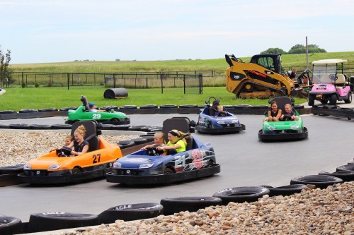 A go-kart track with children racing in colorful karts on a sunny day, surrounded by grass and a construction vehicle.