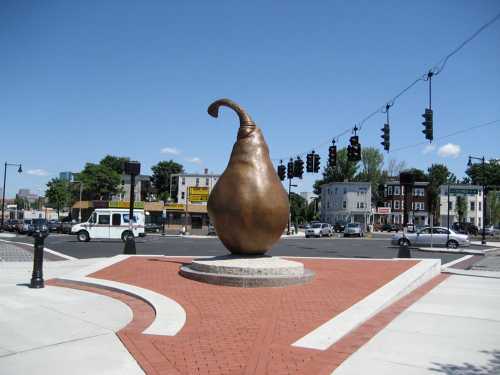 A large bronze pear-shaped sculpture stands in a plaza, surrounded by a road and traffic lights under a clear blue sky.