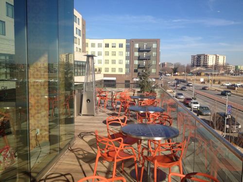 Outdoor seating area with orange chairs and tables, overlooking a busy street and modern buildings under a clear blue sky.