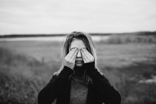 A young woman stands outdoors, covering her eyes with her hands, against a blurred landscape. The image is in black and white.