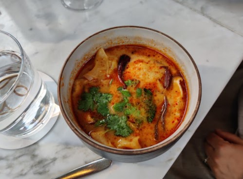 A bowl of spicy soup with dumplings, garnished with cilantro and red chili, on a marble table next to a glass of water.