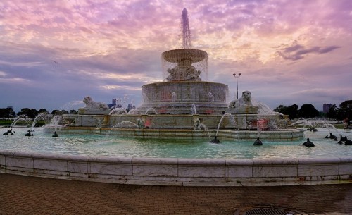 A grand fountain with lion sculptures, surrounded by water, under a colorful sunset sky.