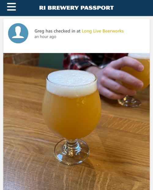 A close-up of a frothy beer in a glass on a wooden table, with a hand in the background.