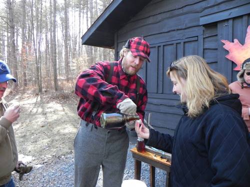 A man in a plaid shirt pours a drink for a woman outside a cabin in a wooded area. Others are gathered nearby.