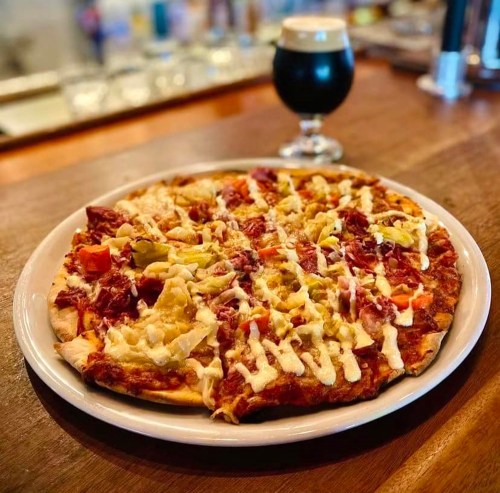 A freshly baked pizza topped with meats, vegetables, and drizzled sauce, served with a dark beer in the background.