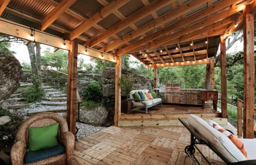 Cozy outdoor patio with wooden beams, string lights, seating area, and a kitchen, surrounded by greenery.