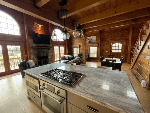 A spacious wooden kitchen with a marble island, gas stove, and a cozy living area featuring large windows and rustic decor.