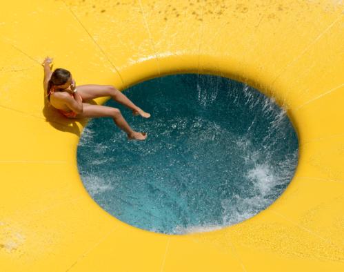 A person slides down a yellow water slide into a blue pool, creating splashes of water.